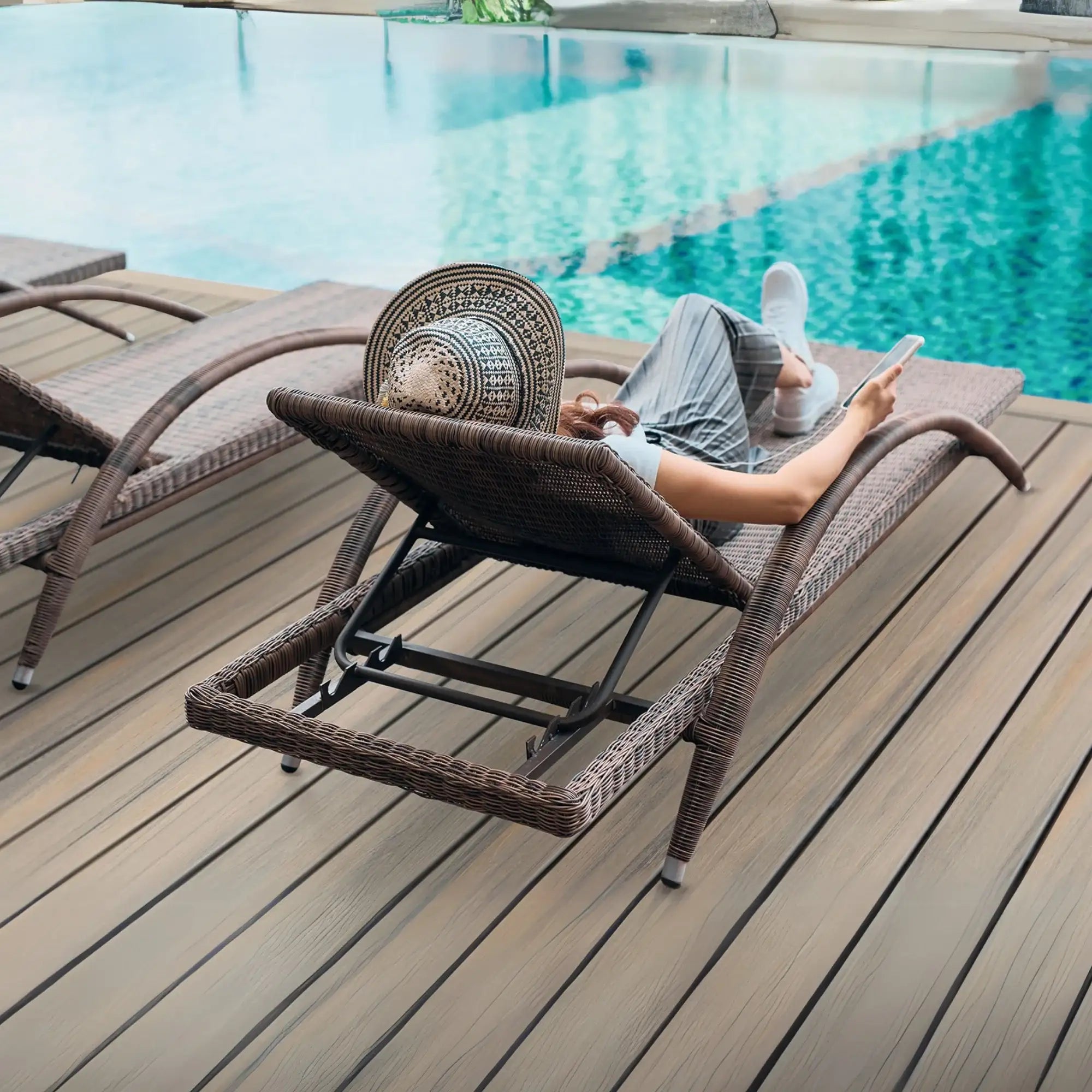 Woman relaxing on wicker lounge chair by pool, using tablet on wooden deck