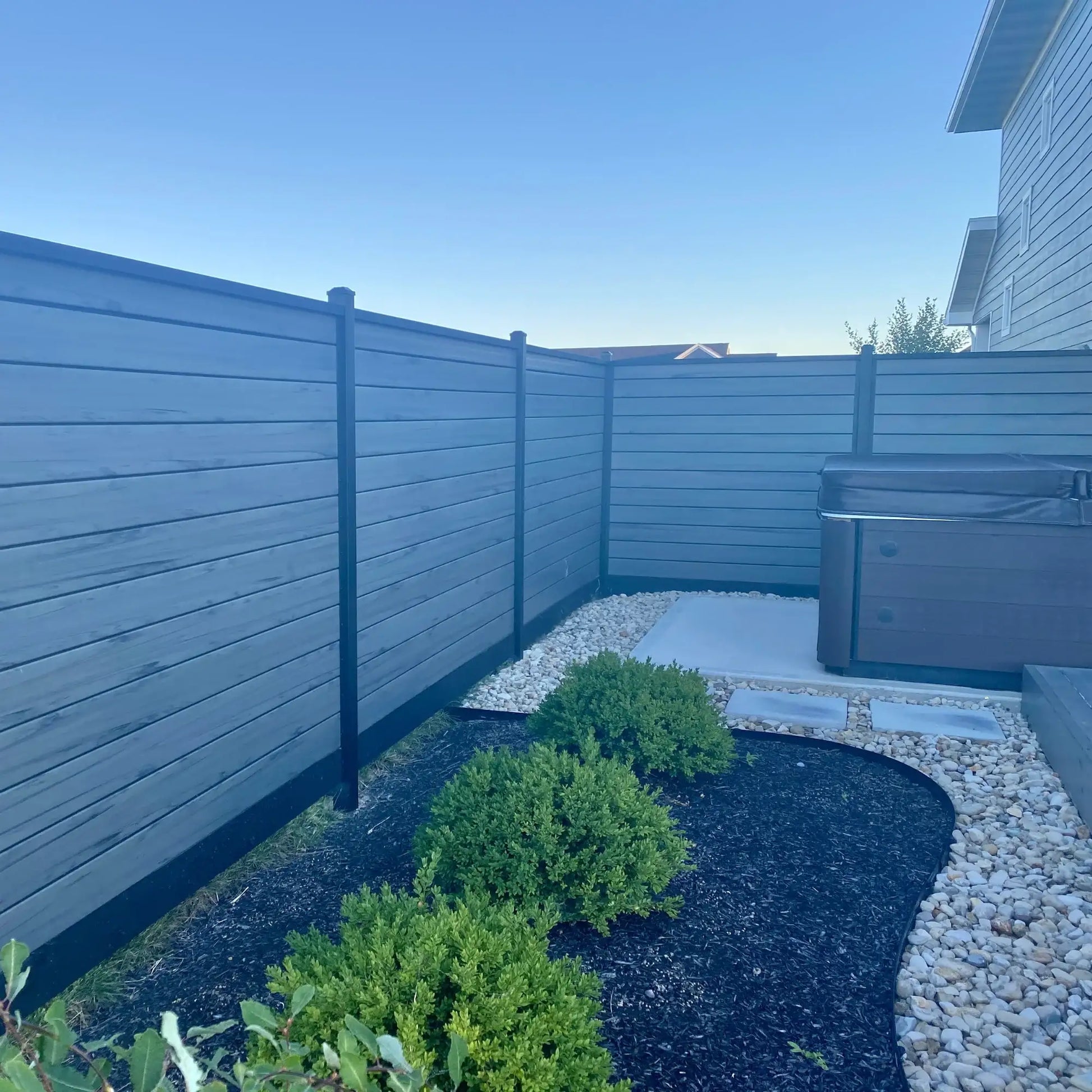 Modern gray horizontal fence with black posts enclosing backyard with hot tub, gravel, and shrubs
