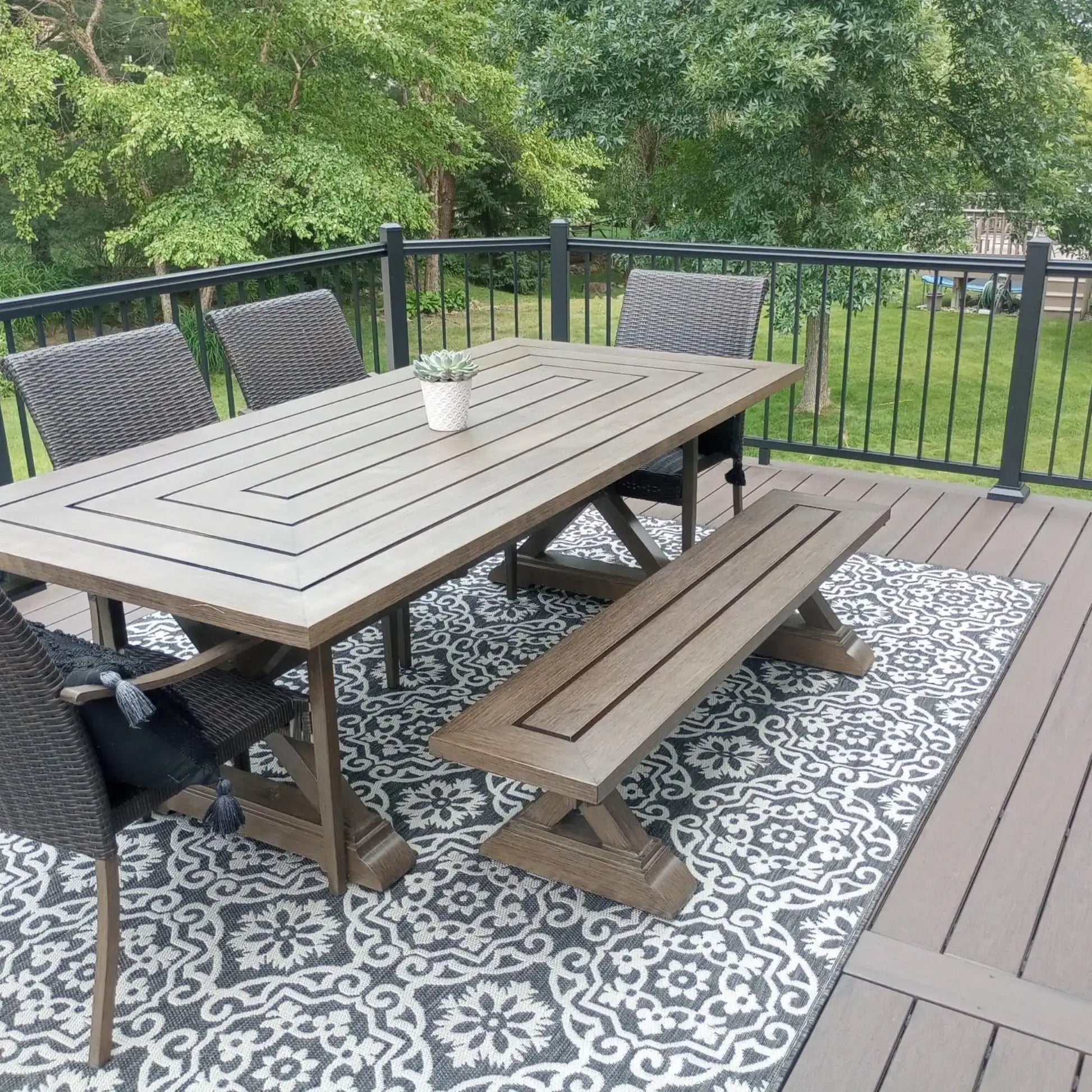 Outdoor dining set with wooden table, bench, wicker chairs on patterned rug, deck with greenery backdrop