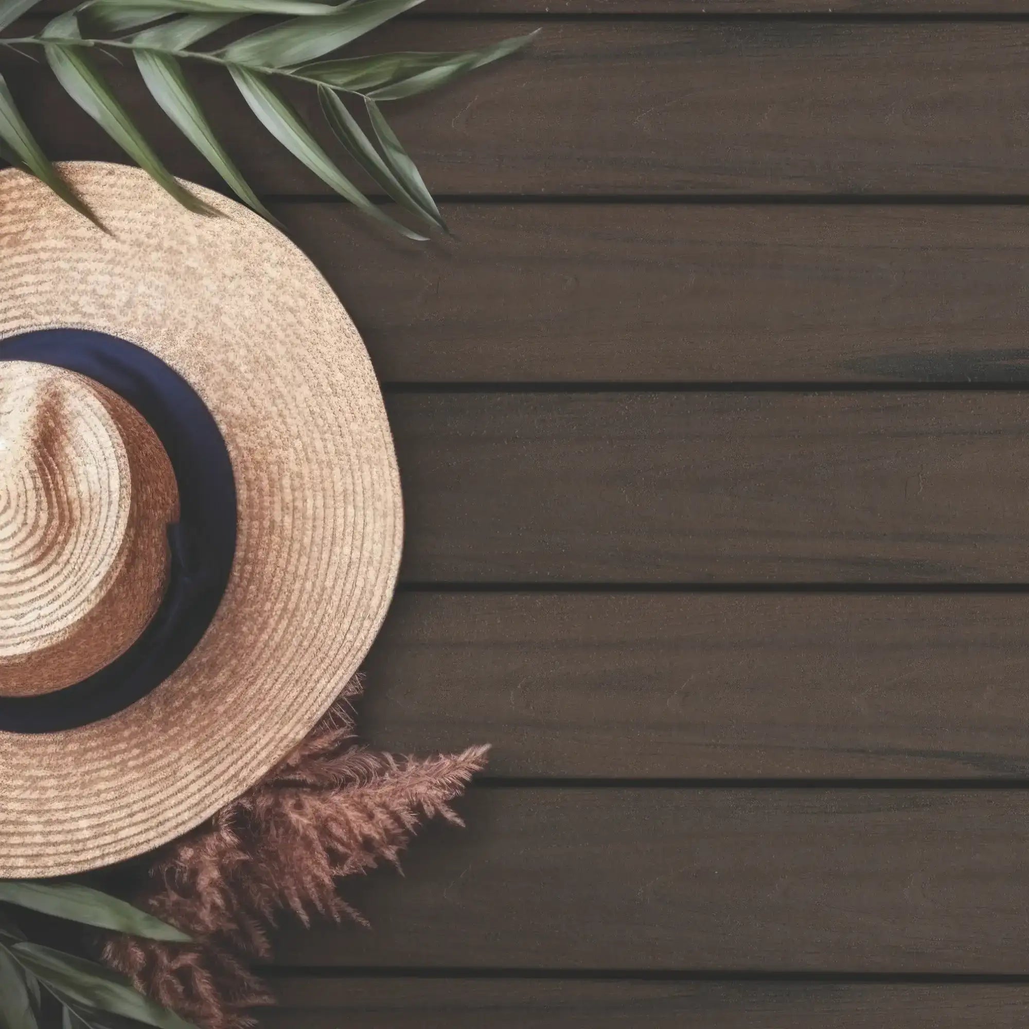 Straw sun hat with navy band on dark wood deck, tropical leaves and pampas grass accent