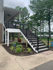 Modern outdoor staircase with black cable railings leading to a covered porch, surrounded by landscaping and lake view