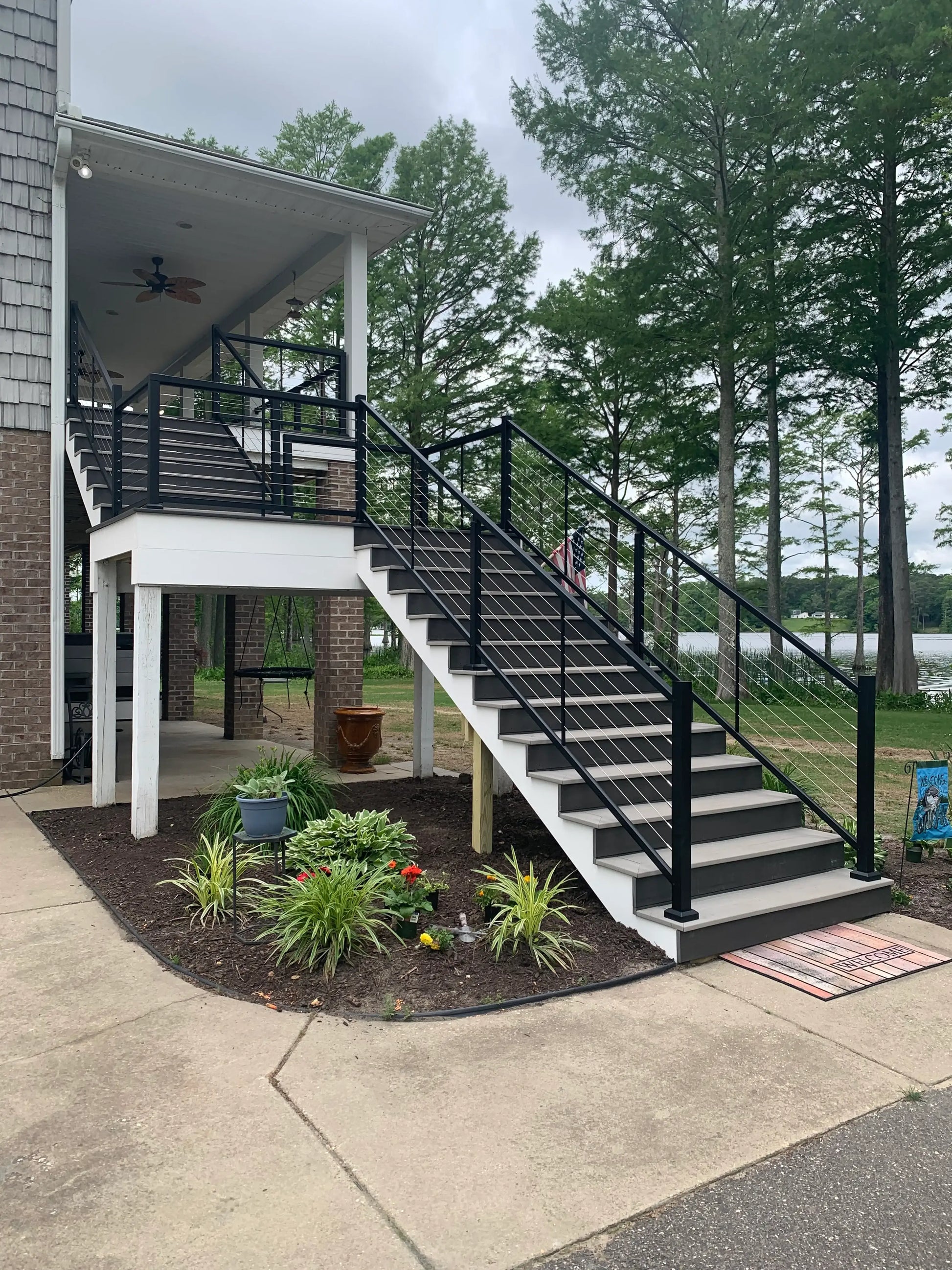 Modern outdoor staircase with black cable railings leading to a covered porch, surrounded by landscaping and lake view