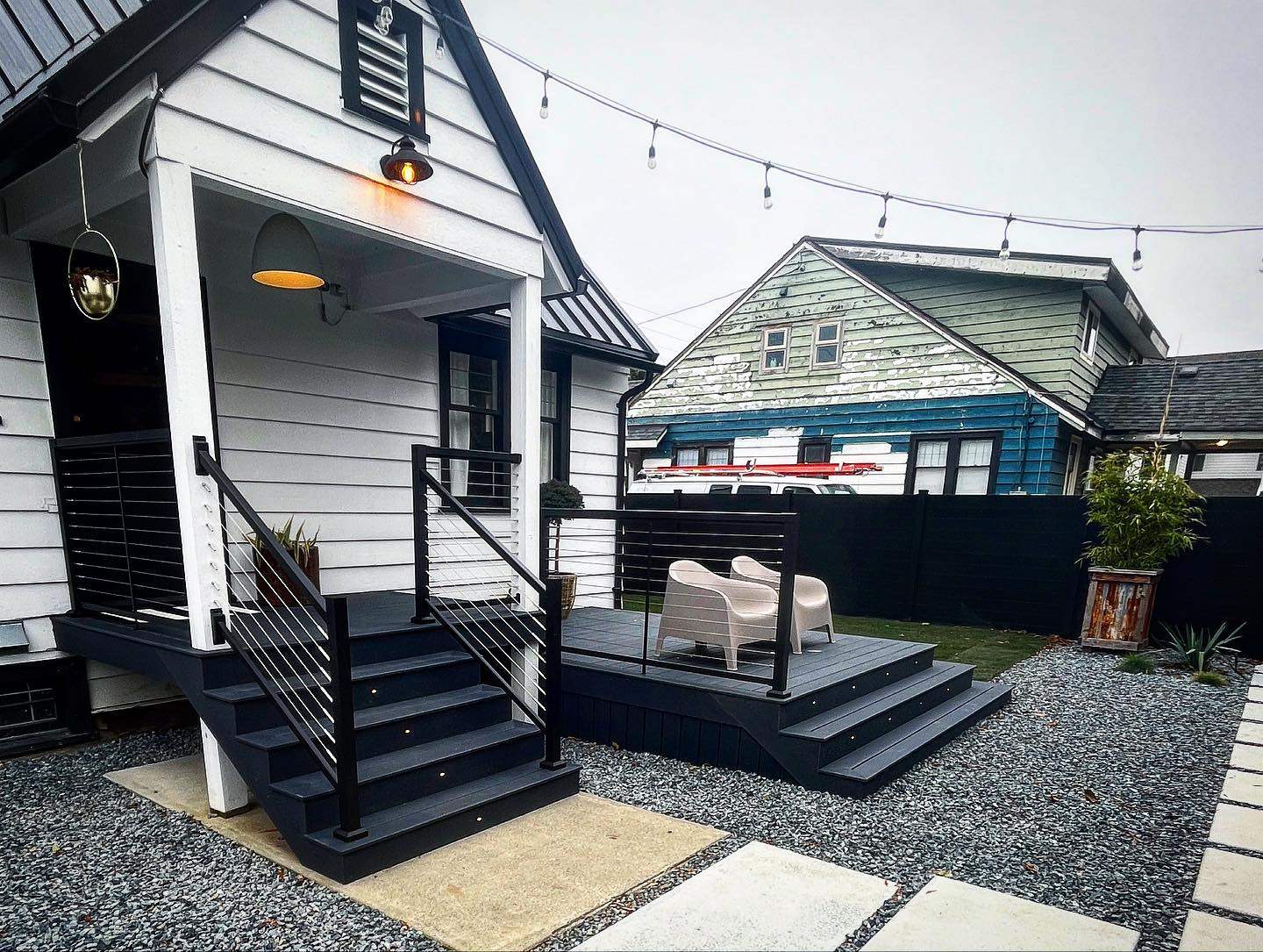 Modern black deck with string lights, outdoor chairs, and gravel yard beside white house