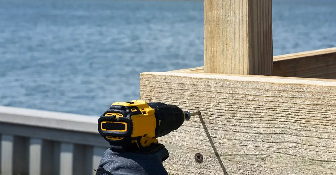 Yellow cordless drill driving screw into wooden deck railing near water, ideal for outdoor construction projects