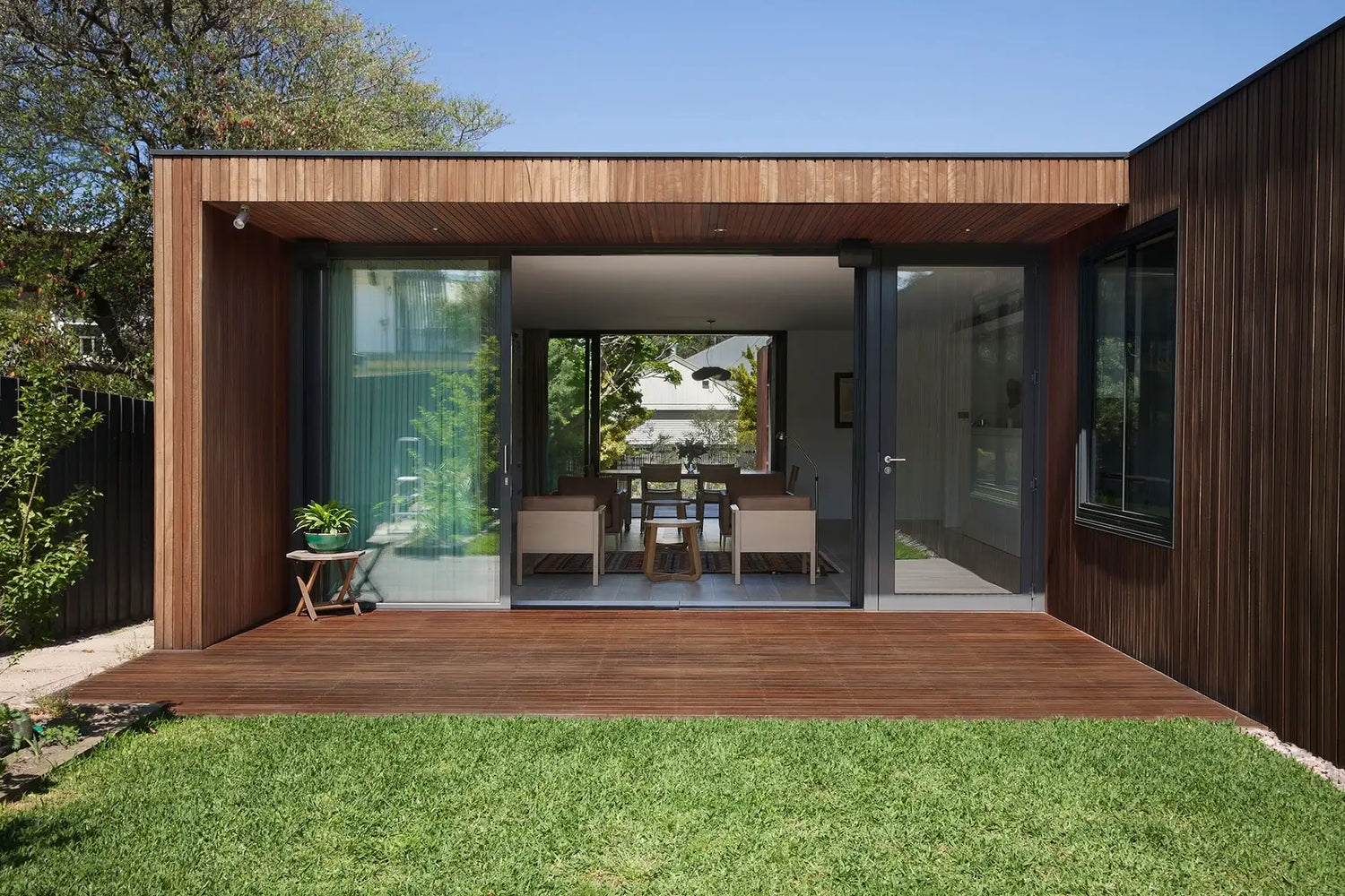 Modern wooden deck with large glass doors opening to indoor dining area, surrounded by green lawn and trees