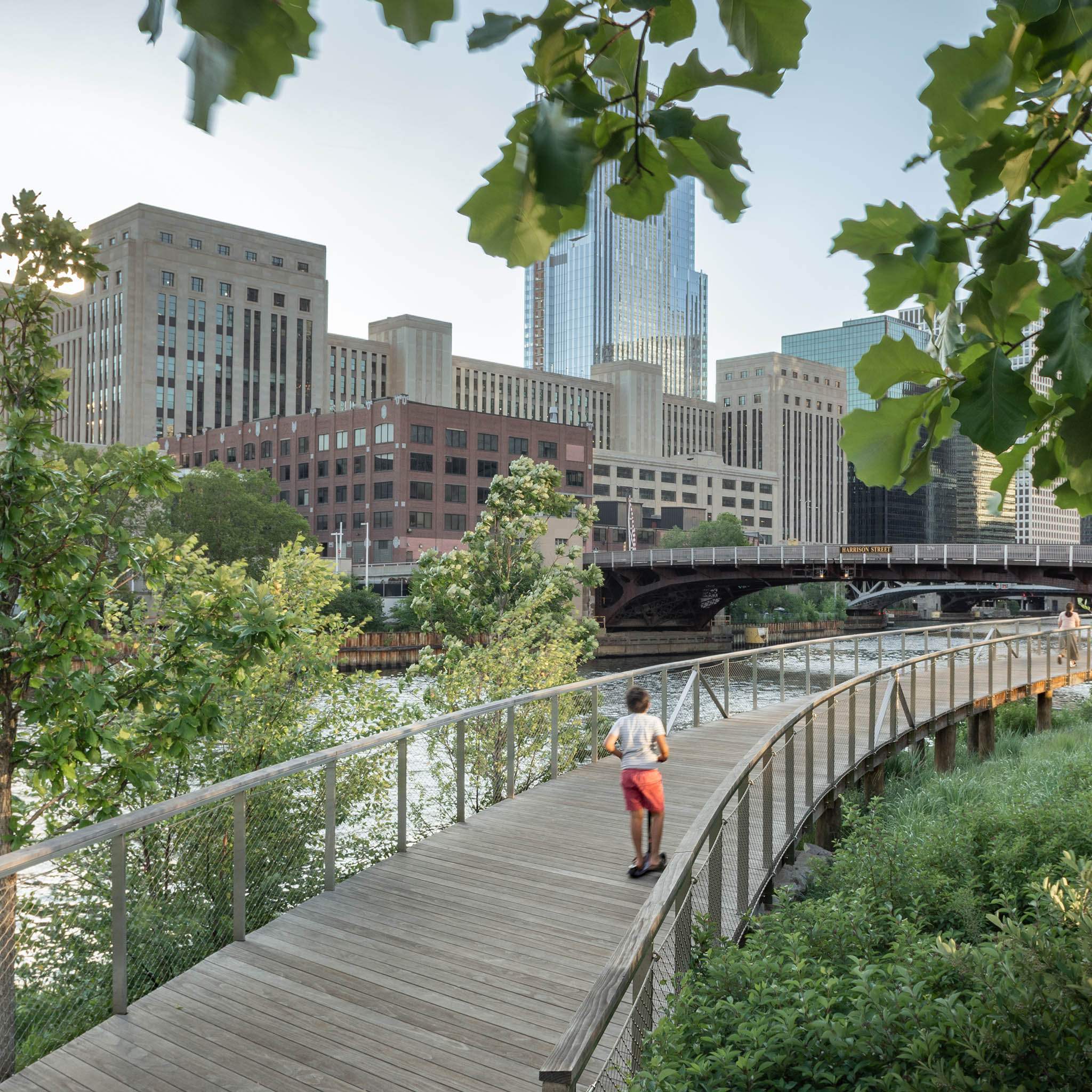 Child riding scooter on wooden riverwalk path with city skyline and Harrison Street bridge in background
