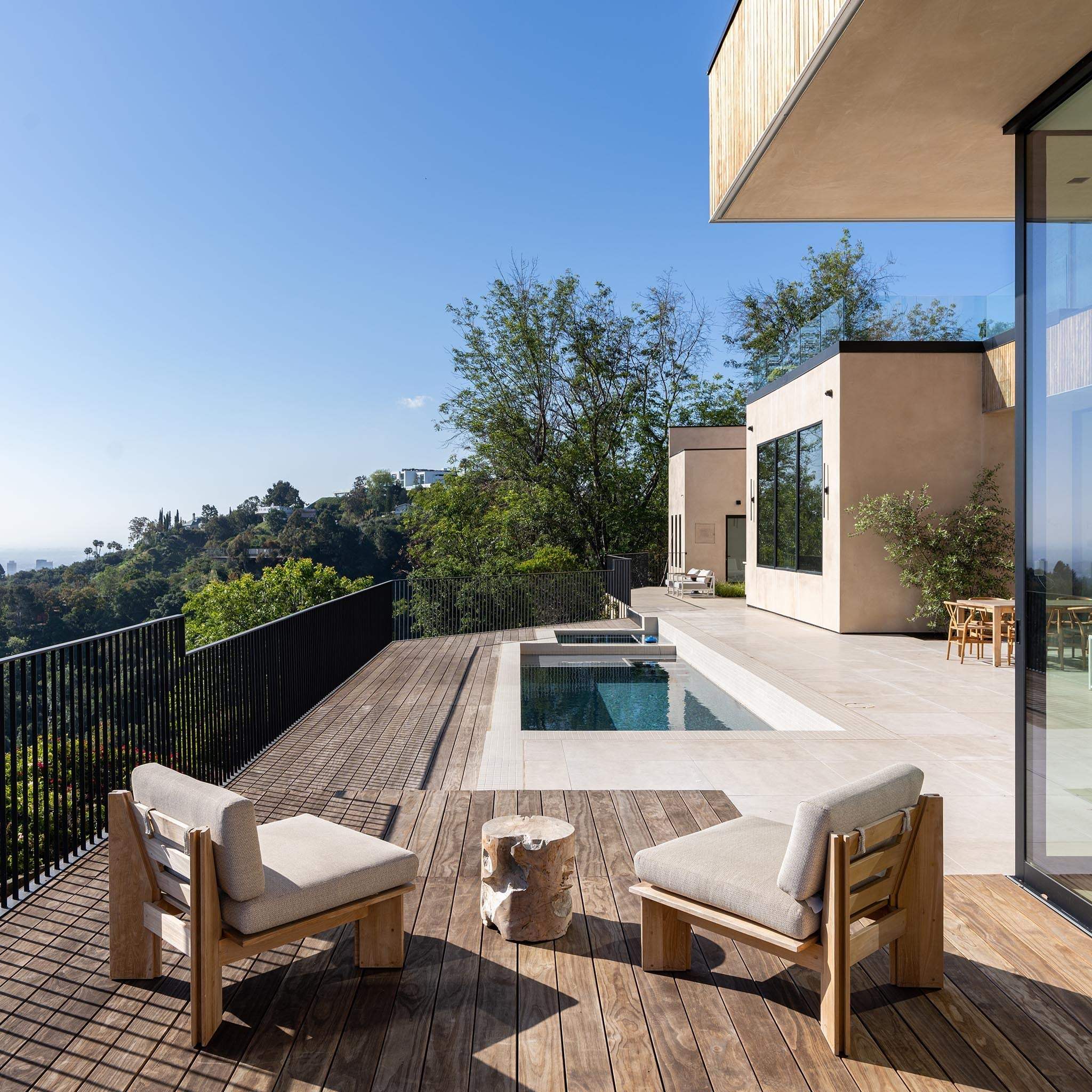 Modern outdoor patio with wooden deck, lounge chairs, and pool overlooking hills under blue sky